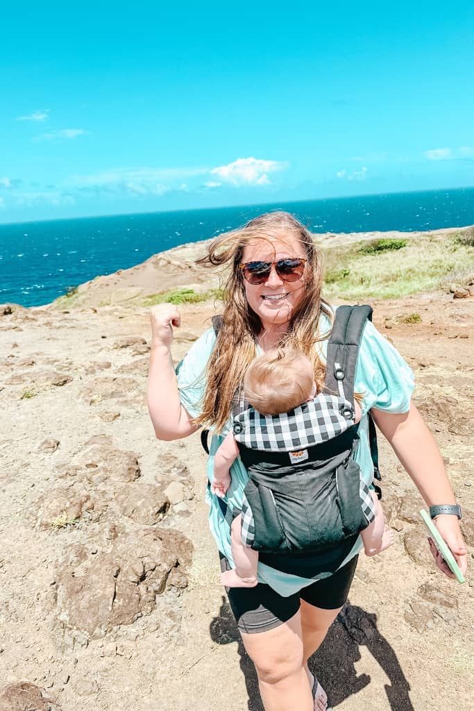 mom holding baby in a baby carrier at the hawaii shoreline with rugged volcanic coastline meeting the ocean on a sunny day 