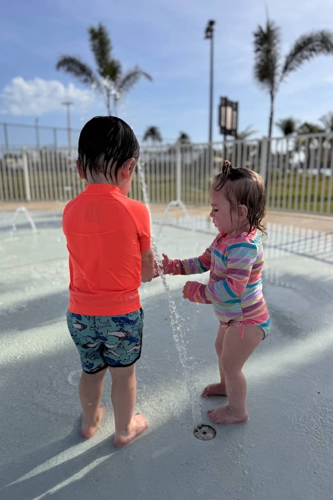 two toddlers playing at a splash pad with baseball fields in the background