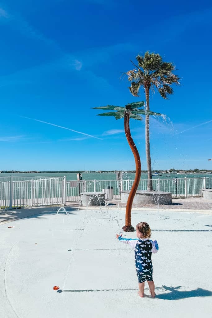 toddler playing at a splash pad with palm trees and water view in the background