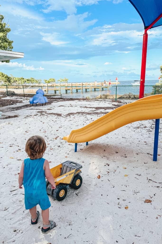 toddler boy playing with a truck in the sand at a park with water in the background and a yellow slide also in the background