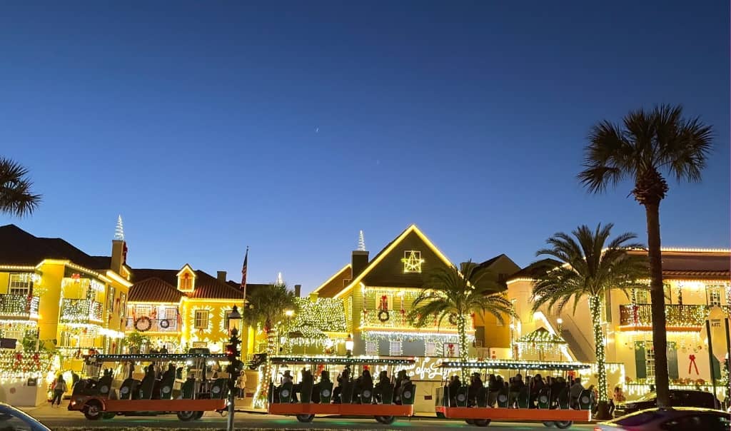 several historic buildings along St. Augustine's waterfront lit up with tons of white christmas tree lights with a trolley in front of it and palm trees on the side