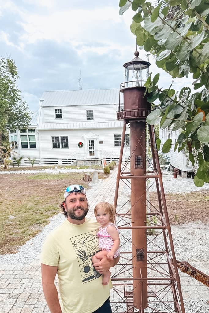 dad holding toddler girl in front of a white historic building and a small model of the sanibel lighthouse