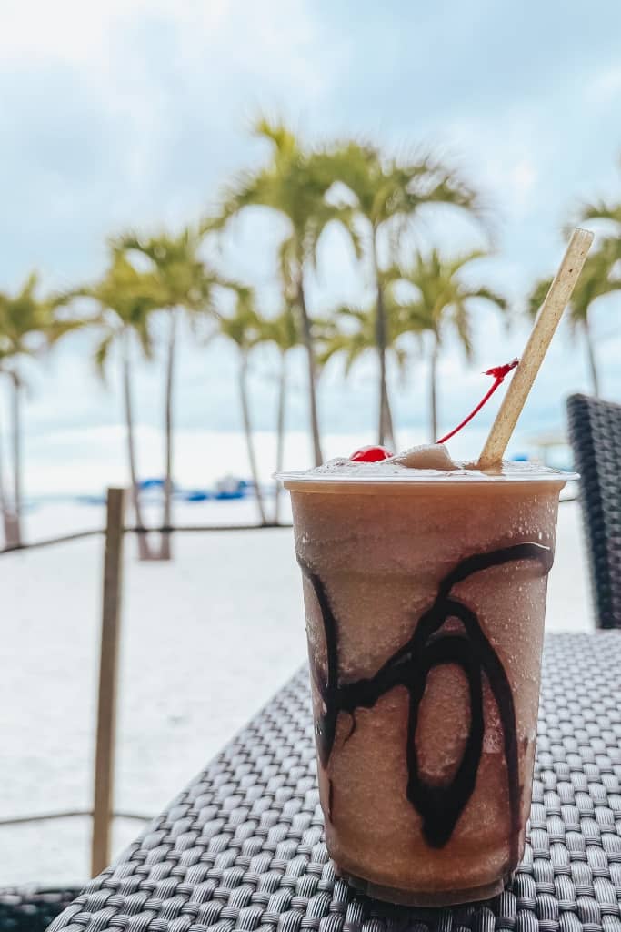 chocolate frozen cocktail drink on the table with palm trees and the beach in the background at a beach bar