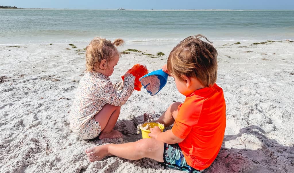 two toddlers sitting in the sand playing with sand toys at sand key with the florida beach in the background
