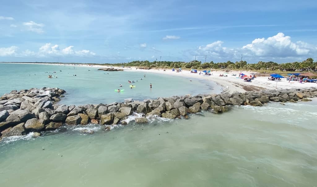 view of a beach at fort desoto from the pier with rocks along the side of it and tons of beachgoers in the water and laying out on beach chairs