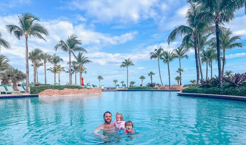 dad holding 1 year old and 2 year old in the swimming pool on a sunny day with tons of palm trees