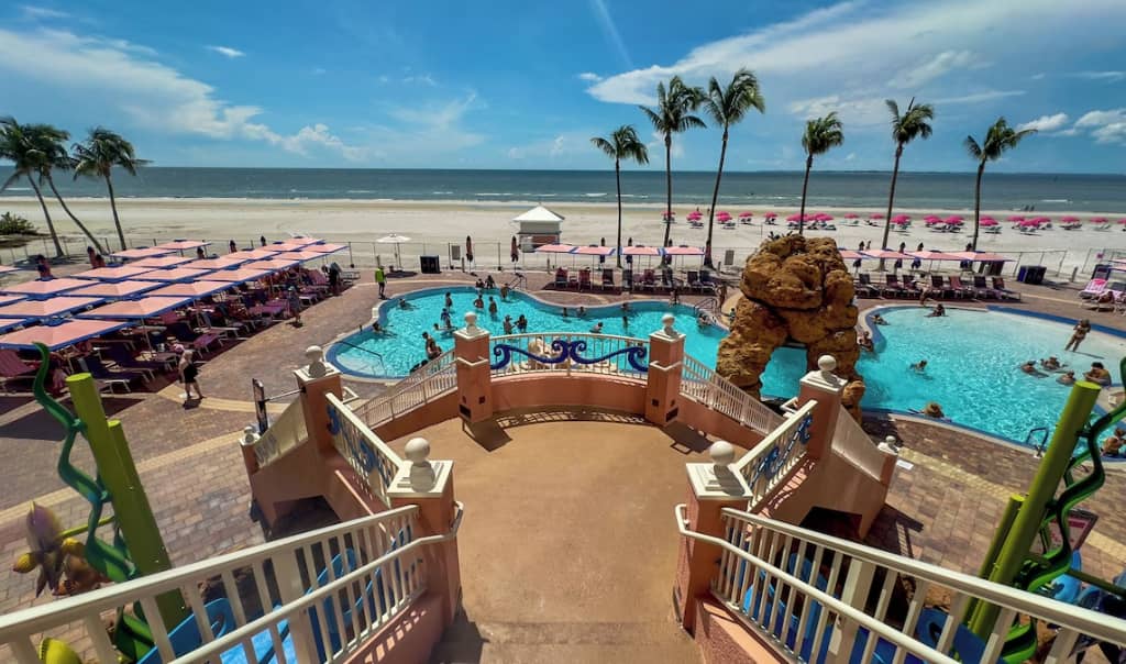photo of large pool surrounded by palm trees on the beach taken from the top of grand steps leading down to the pool's courtyard