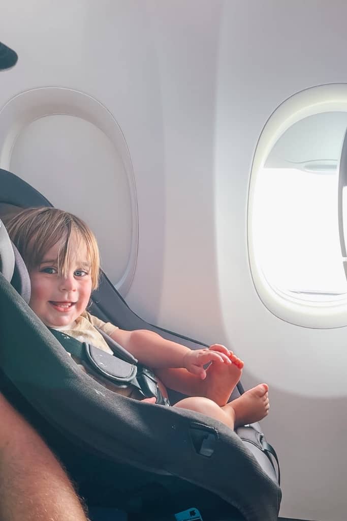 toddler boy sitting in his car seat on the airplane and smiling at the camera 