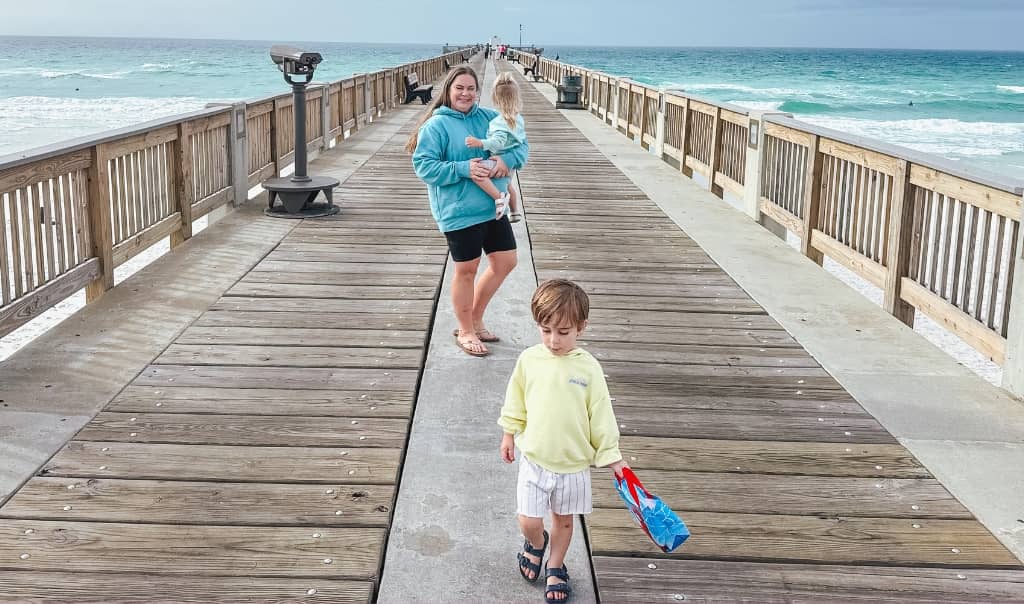 mom holding toddler while another toddler runs along a pier extending into the ocean on a cloudy day