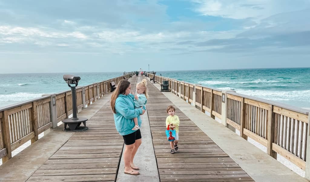 mom holding toddler while another toddler runs along a pier extending into the ocean on a cloudy day