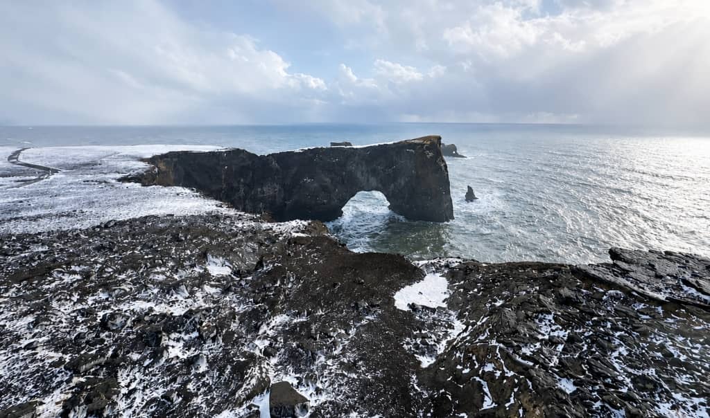 towering cliff rising out of the sea with a natural arch cut out of it and waves crashing along the cliff on a winter day with snow on the ground in iceland