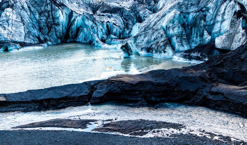 A bright blue glacier flows between rugged mountains, with snow-covered peaks and rocky terrain surrounding the ice
