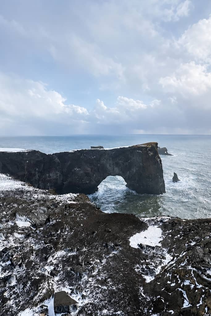towering cliff rising out of the sea with a natural arch cut out of it and waves crashing along the cliff on a winter day with snow on the ground in iceland