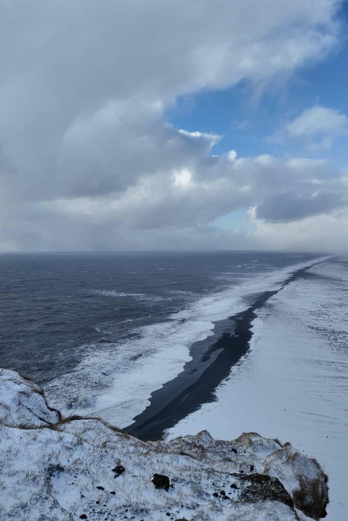 view of a black sand beach taken from the top of a cliff with rocky edges in the frame, shoreline covered in snow but you can see the black sand beach where the waves crash to shore
