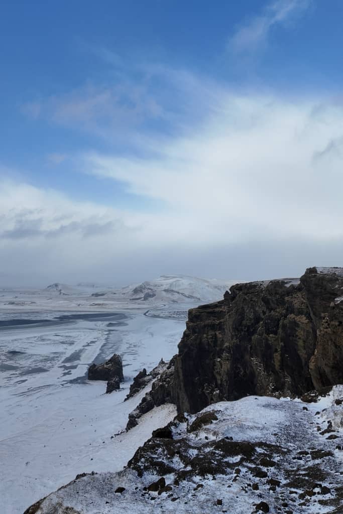 views of dramatic tall cliffs meeting the sea with a black sand beach lining the sea covered in snow