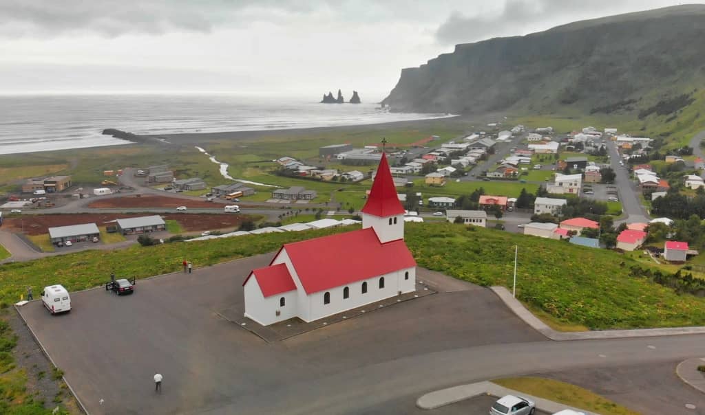 white church with red roof and steeple on a mountain overlooking a village town, the ocean, and large cliffs