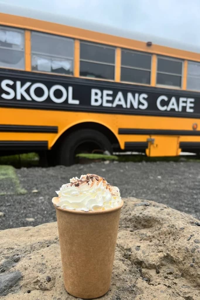 yellow school bus with a sign that says skool beans cafe written in white with a black background and a coffee topped with whipped cream sitting on top of a table in front of the school bus